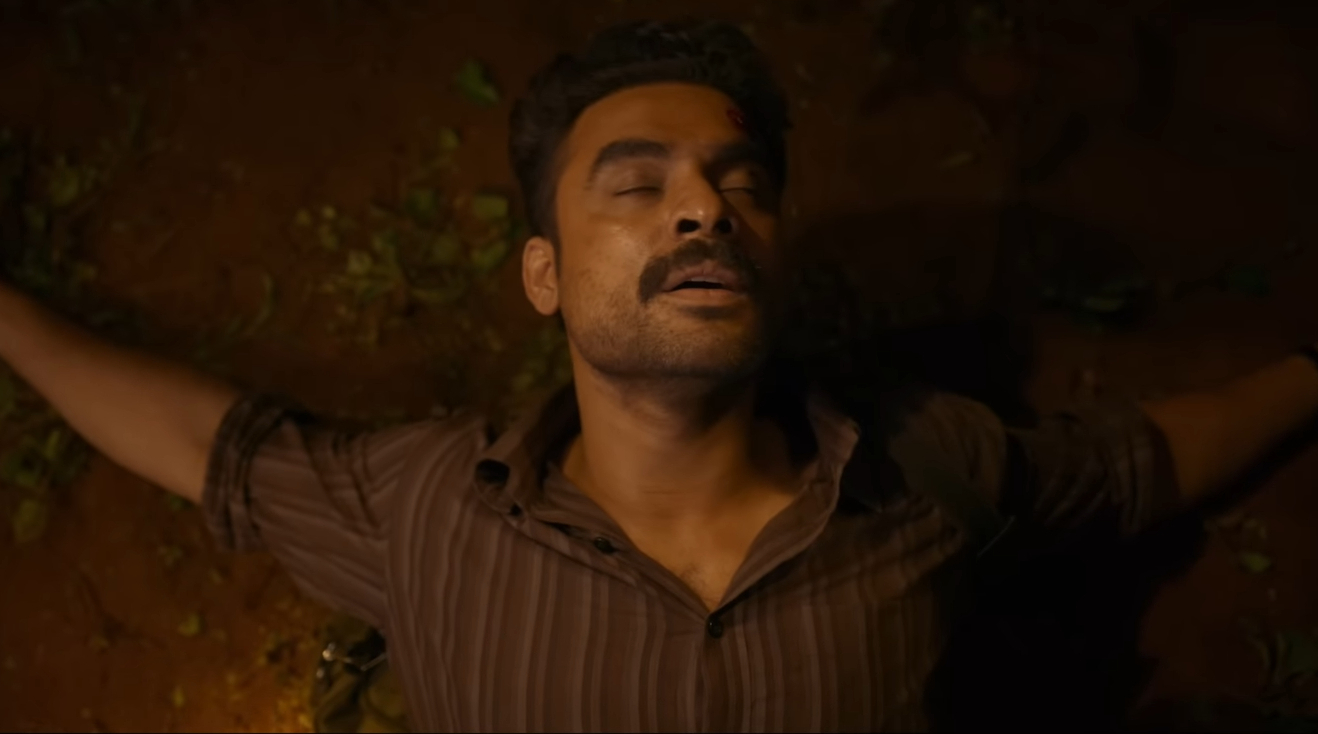 A man lying on the ground with a contemplative expression, surrounded by earthy tones and foliage.