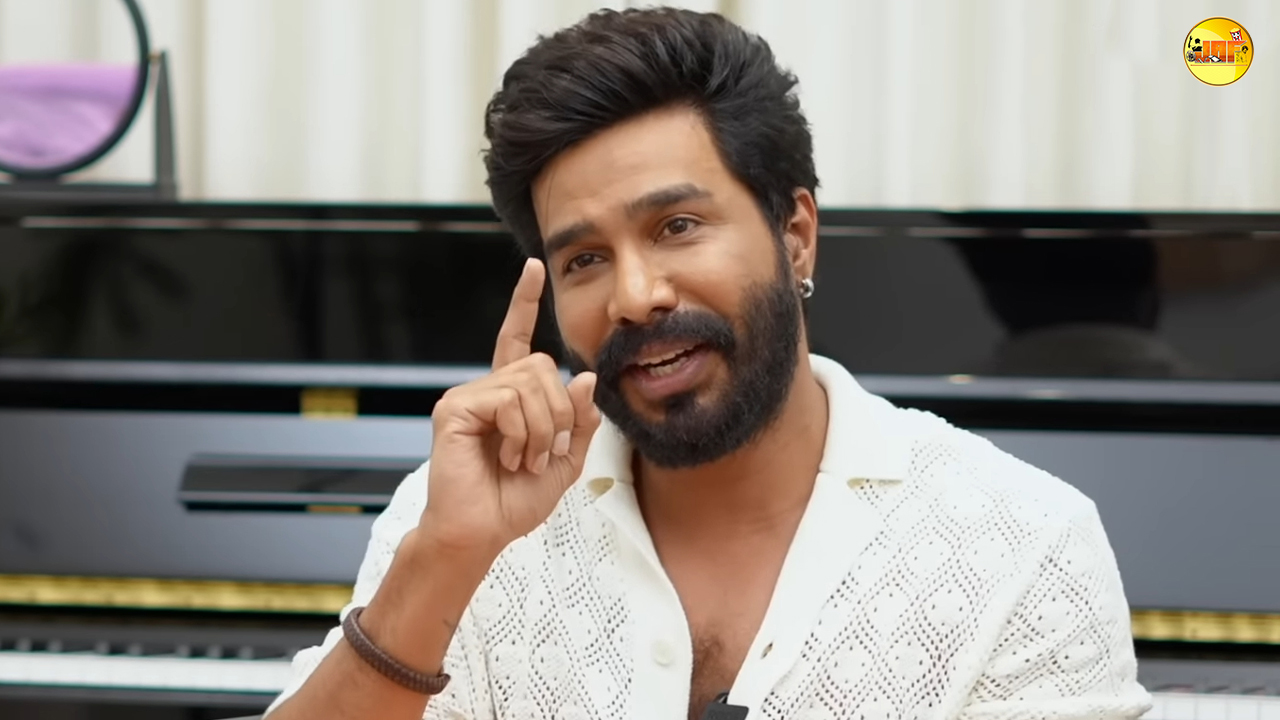 A well-groomed man with a beard gestures expressively during a conversation, seated in front of a piano, conveying an enthusiastic demeanor.